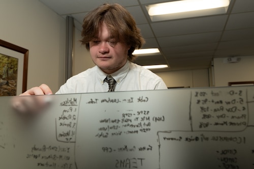 A man stands behind a clear writing board, taking notes.