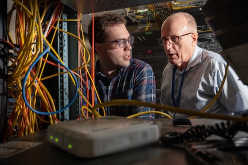 Two men are standing and talking, framed by an opening in a server rack, where electronic devises and wires can be seen.