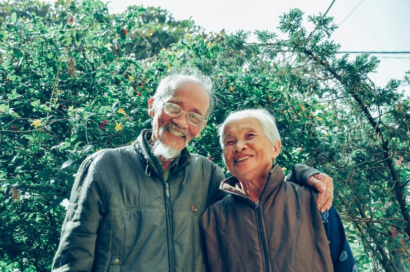 Elderly man and woman smile together in front of greenery