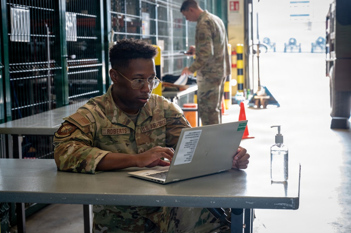 A man in a camouflage military uniform sits at a table looking at a computer screen while in a warehouse. Another man in similar attire stands in the background, holding a phone.