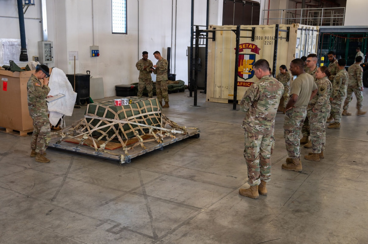 Several people in camouflage military uniforms stand in a warehouse looking at a cargo pallet on the floor. There is a large container and workout equipment in the background.