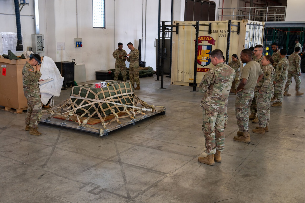 Several people in camouflage military uniforms stand in a warehouse looking at a cargo pallet on the floor. There is a large container and workout equipment in the background.