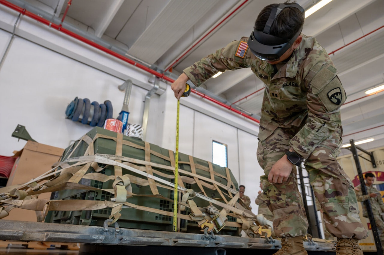A person in a camouflage military uniform bends over to look at a measurement of a cargo pallet while wearing a headset in a warehouse.