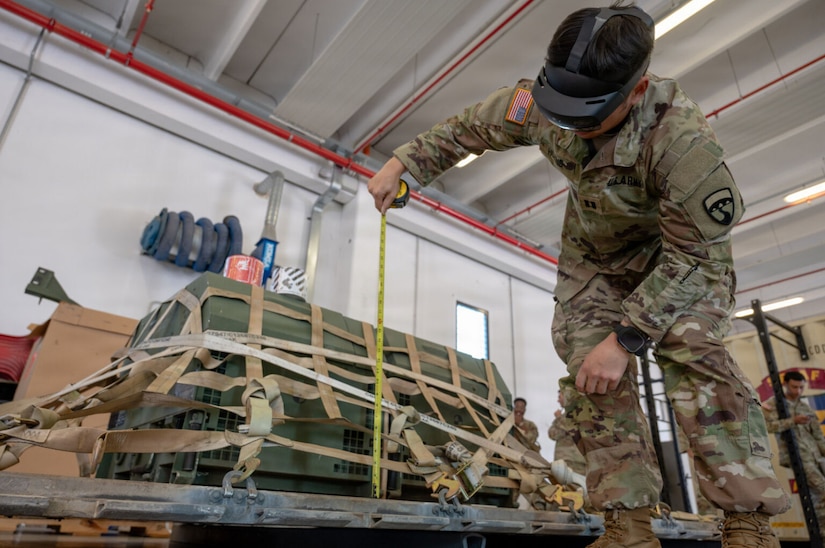 A person in a camouflage military uniform bends over to look at a measurement of a cargo pallet while wearing a headset in a warehouse.