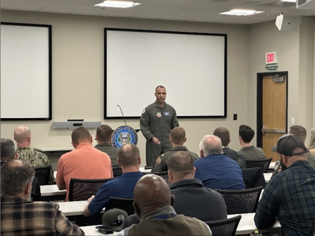 A mean wearing a flight suit stands before a group of people