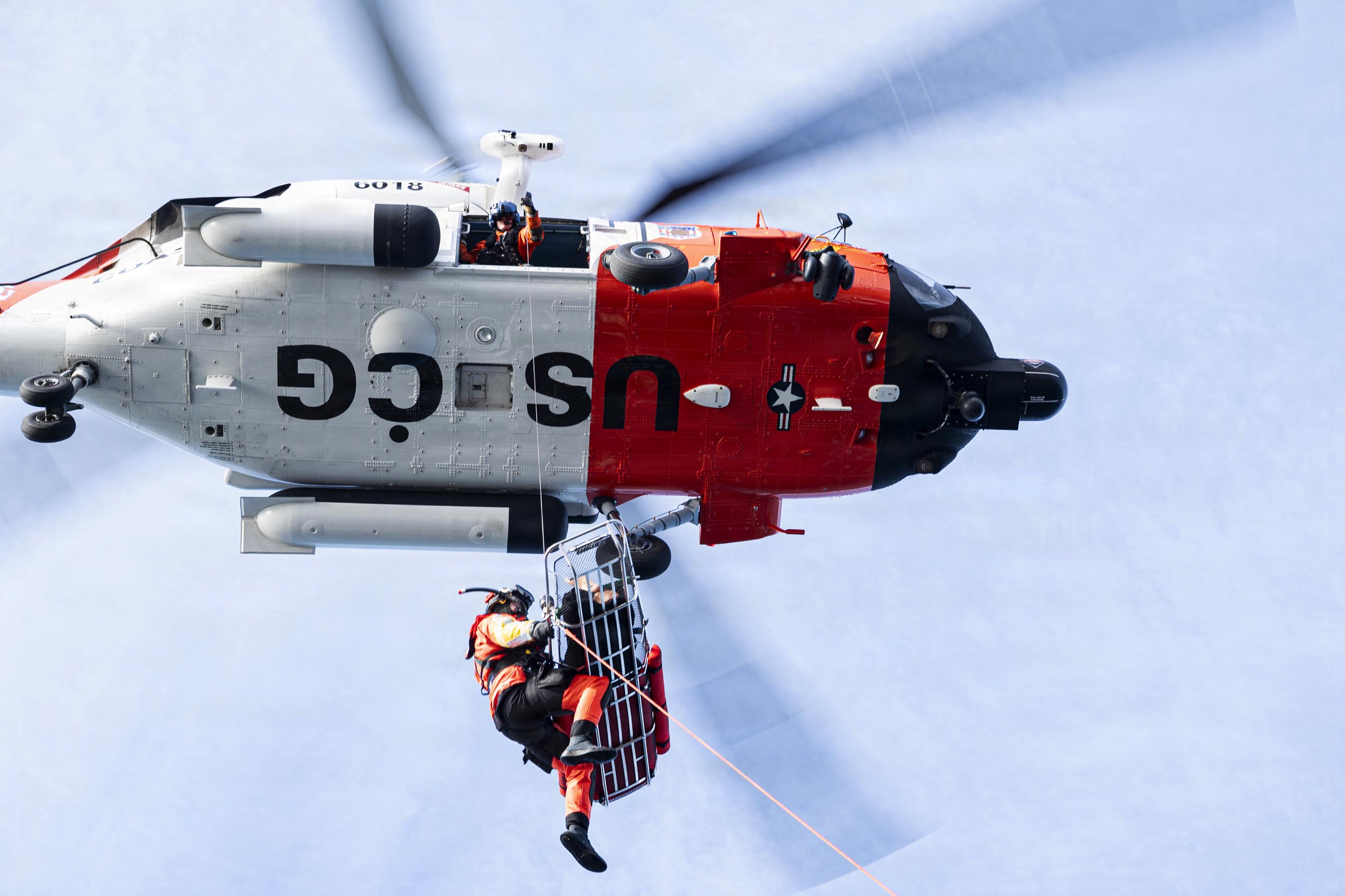 A helicopter hovers in a blue sky with a person at an open door holding a cable that is attached to a stretcher with a person in it and another holding its side.