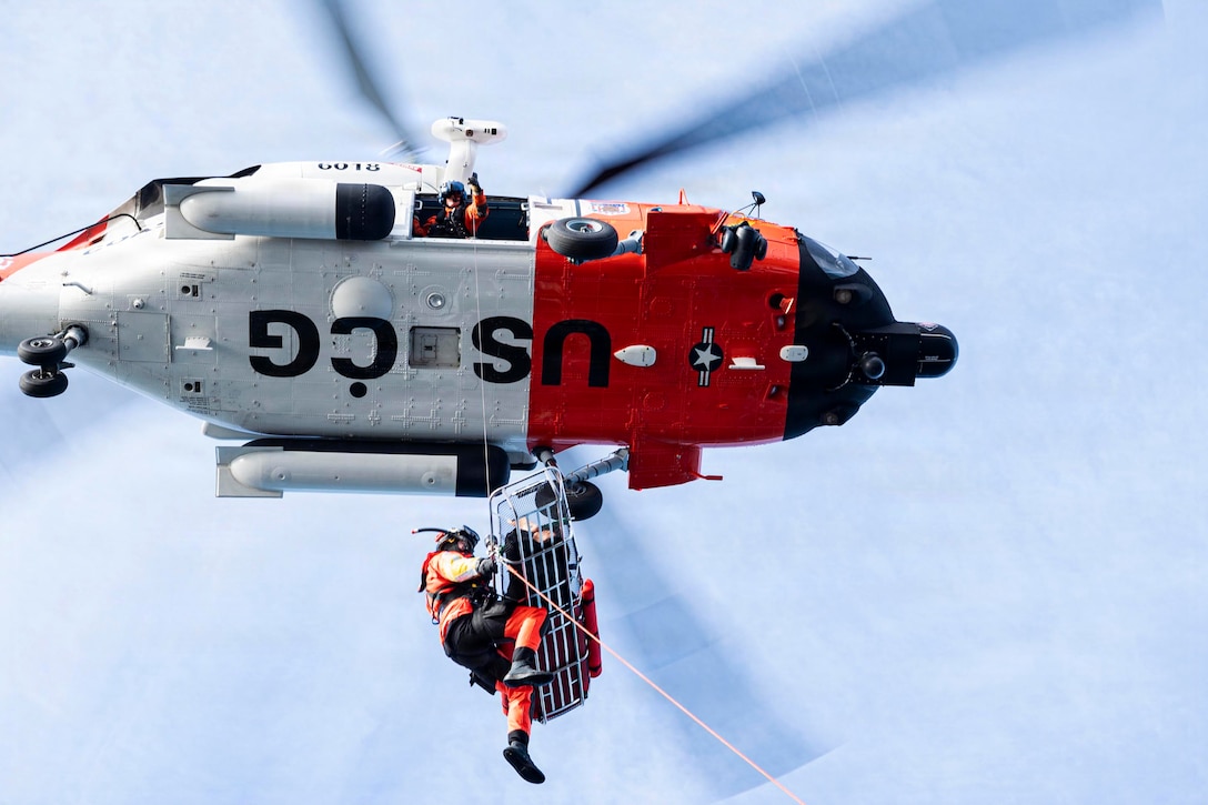 A helicopter hovers in a blue sky, with a person holding a cable from an open door; the cable is attached to a stretcher that has one person in it and another person holding its side.