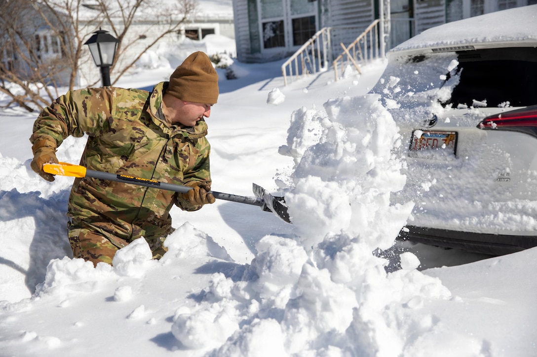 A man wearing a camouflage uniform, hat and gloves shovels thigh-level snow from behind a car in a driveway. There are two houses in the background.