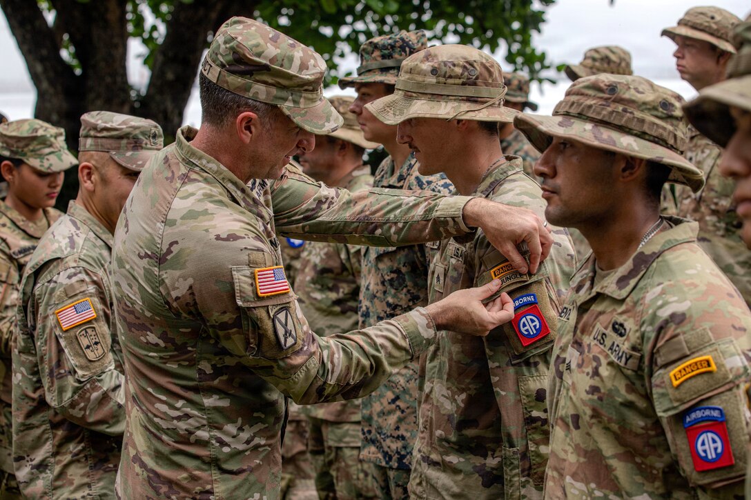 A soldier puts a tab on the sleeve of another soldier's uniform as others stand nearby.
