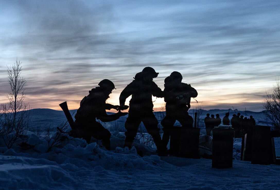Three Marines in silhouette, one carrying a mortar, move on hilly terrain against a dusky light-streaked sky.
