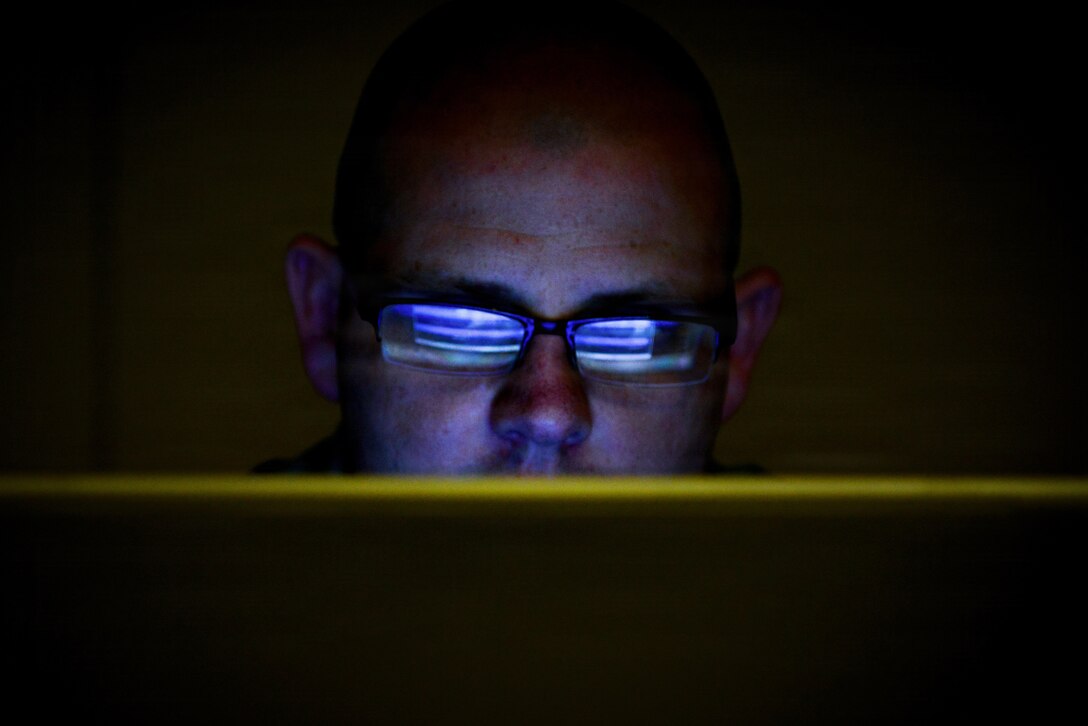 Lines of blue light are reflected in the eyeglasses of a service member looking an a screen in the dark.