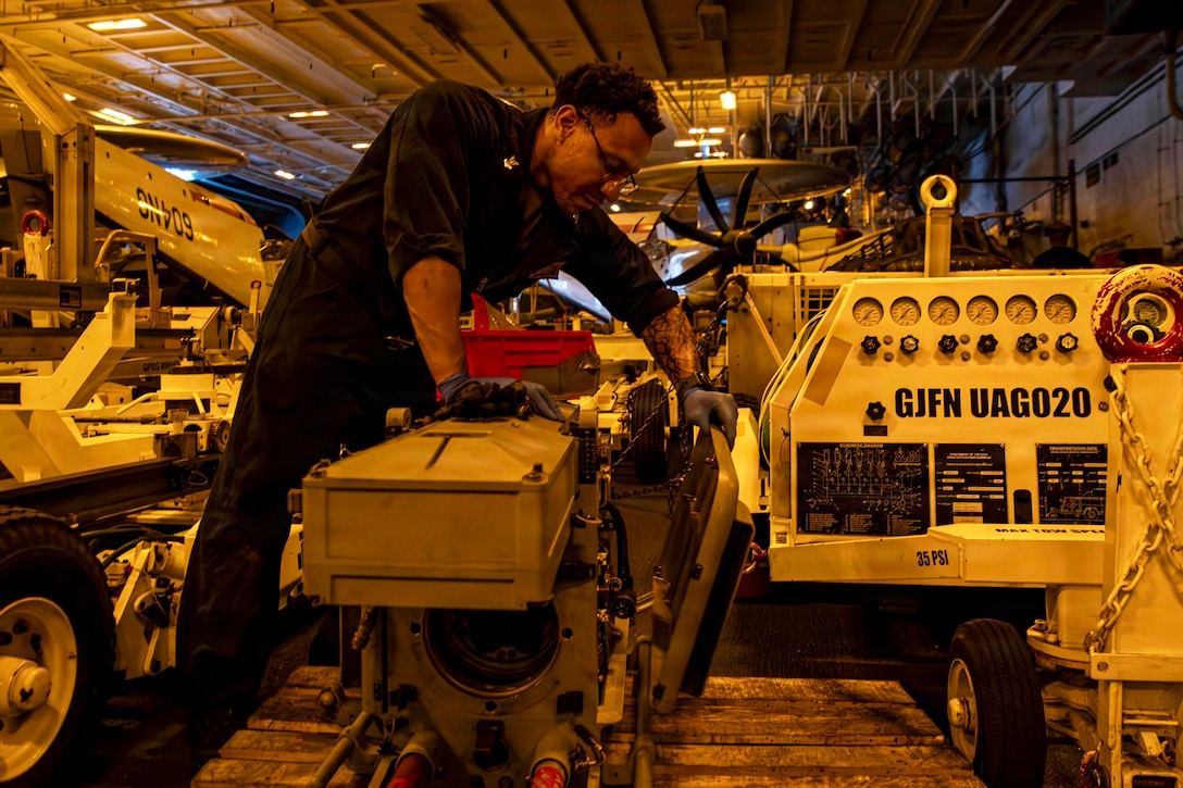 A man wearing coveralls, glasses and gloves in a dimly lit area looks down while opening the side panel on a piece of equipment. There is more equipment scattered nearby.