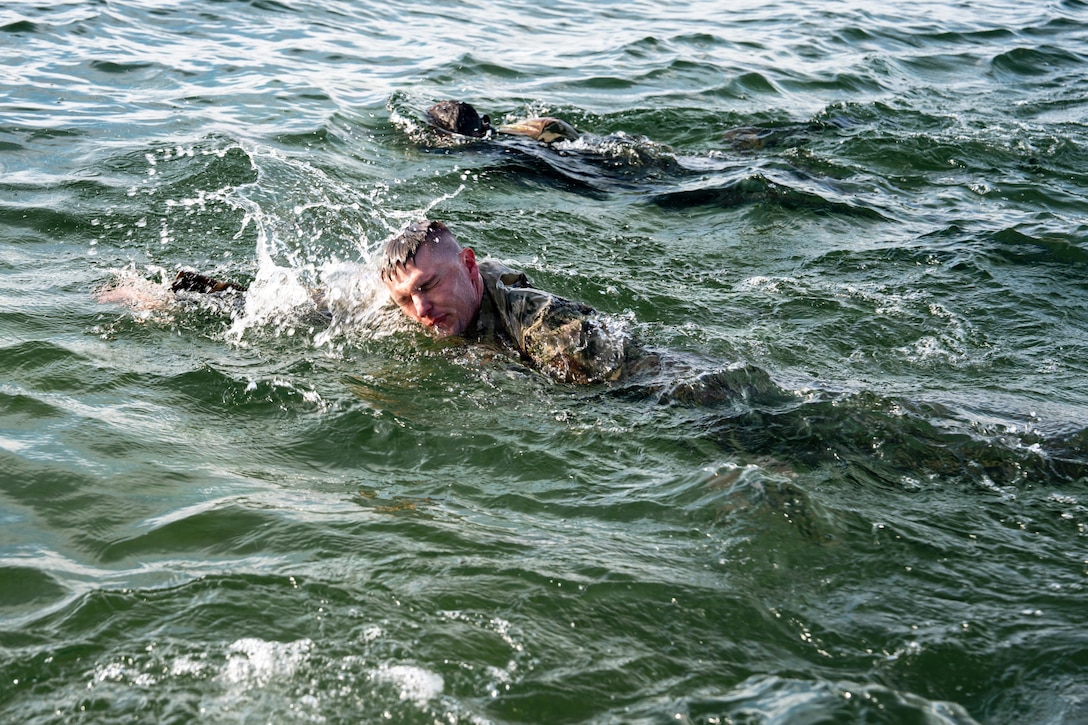 A man in a camouflage military uniform swims through the water, with another person in similar attire doing the same nearby.