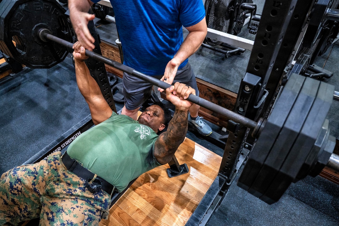 A person wearing a green T-shirt and camouflage pants lies on an exercise bench while preparing to bench press large weights. Another person in athletic attire is standing behind him, ready to assist.