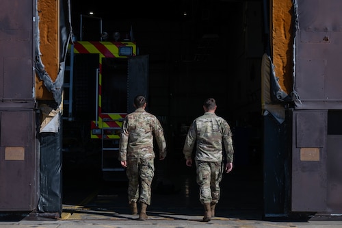 2 men walk into a hangar. There is a firetruck inside of the hangar on the left. The inside looks dramatically darker than the outside.