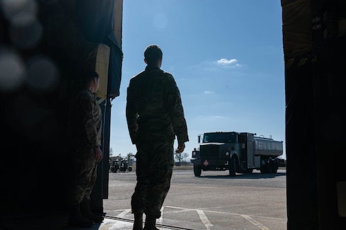 2 men standing at the large doors of a hangar. They are watching outside as a refueler approaches them.