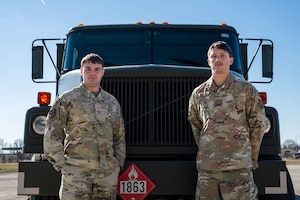 2 men standing in front of a large, green, refueler truck. It is a bright, blue day. The man standing on the left has his hands in his pockets, as the man on the right is standing with his hands behind his back. Both men are in camouflage uniforms.
