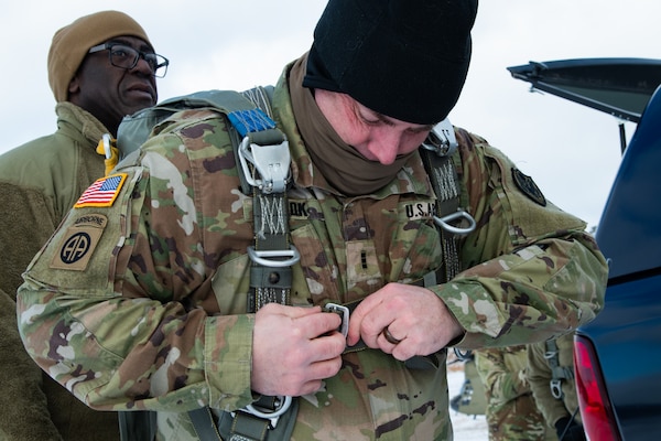 A man wearing a camouflage military uniform prepares for a parachute jump outside in a snow covered field.