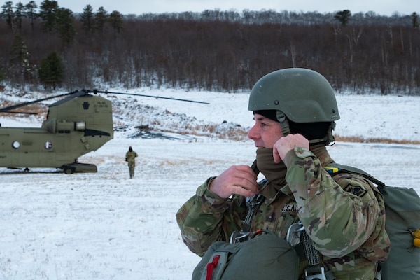 A man wearing a camouflage military uniform prepares for a parachute jump outside in a snow covered field.