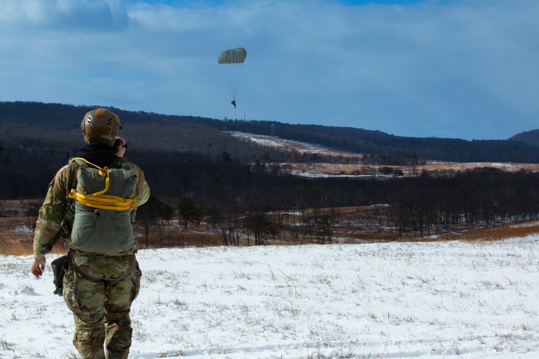 A man wearing a camouflage military uniform prepares for a parachute jump outside in a snow covered field.
