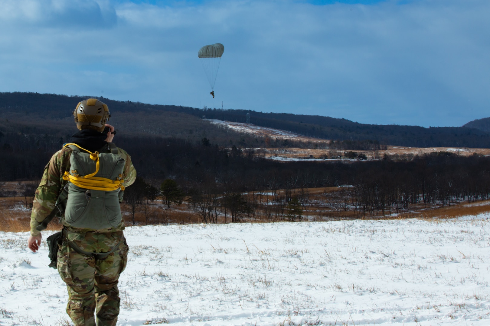 A man wearing a camouflage military uniform prepares for a parachute jump outside in a snow covered field.