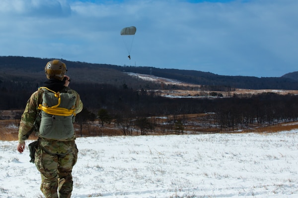 A man wearing a camouflage military uniform prepares for a parachute jump outside in a snow covered field.