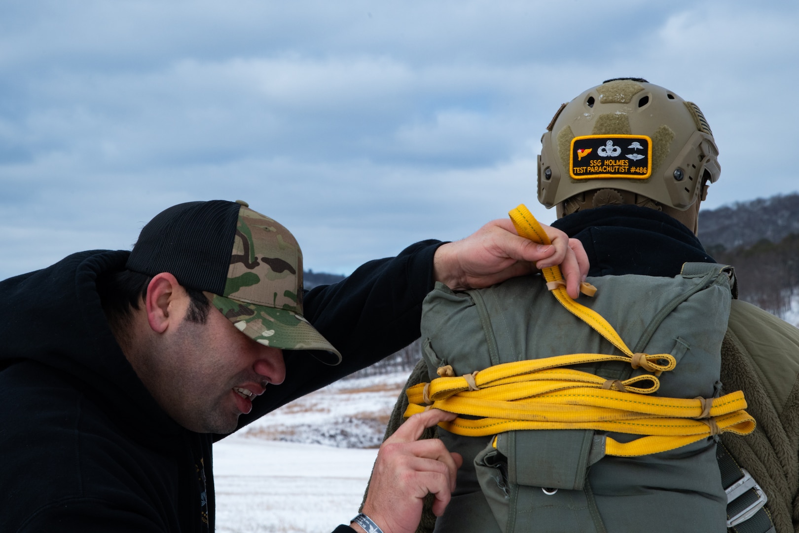 A man wearing a camouflage military uniform prepares for a parachute jump outside in a snow covered field.