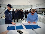 Lt. Cmdr. Brendan Kelley (left), commanding officer of USCGC Vincent Danz (WPC 1162), looks on as Allen Harker (right), contracting officer for the Fast Response Cutter Project, signs the acceptance of the cutter during a ceremony in Key West, Florida, on Feb. 26, 2026. The cutter is named for Vincent Danz, a New York City police officer and U.S. Coast Guard reservist who made the ultimate sacrifice on Sept. 11, 2001, while responding to the attacks on the World Trade Center. (U.S. Coast Guard photo)