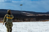 A man wearing a camouflage military uniform prepares for a parachute jump outside in a snow covered field.