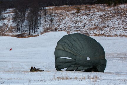 A man wearing a camouflage military uniform prepares for a parachute jump outside in a snow covered field.