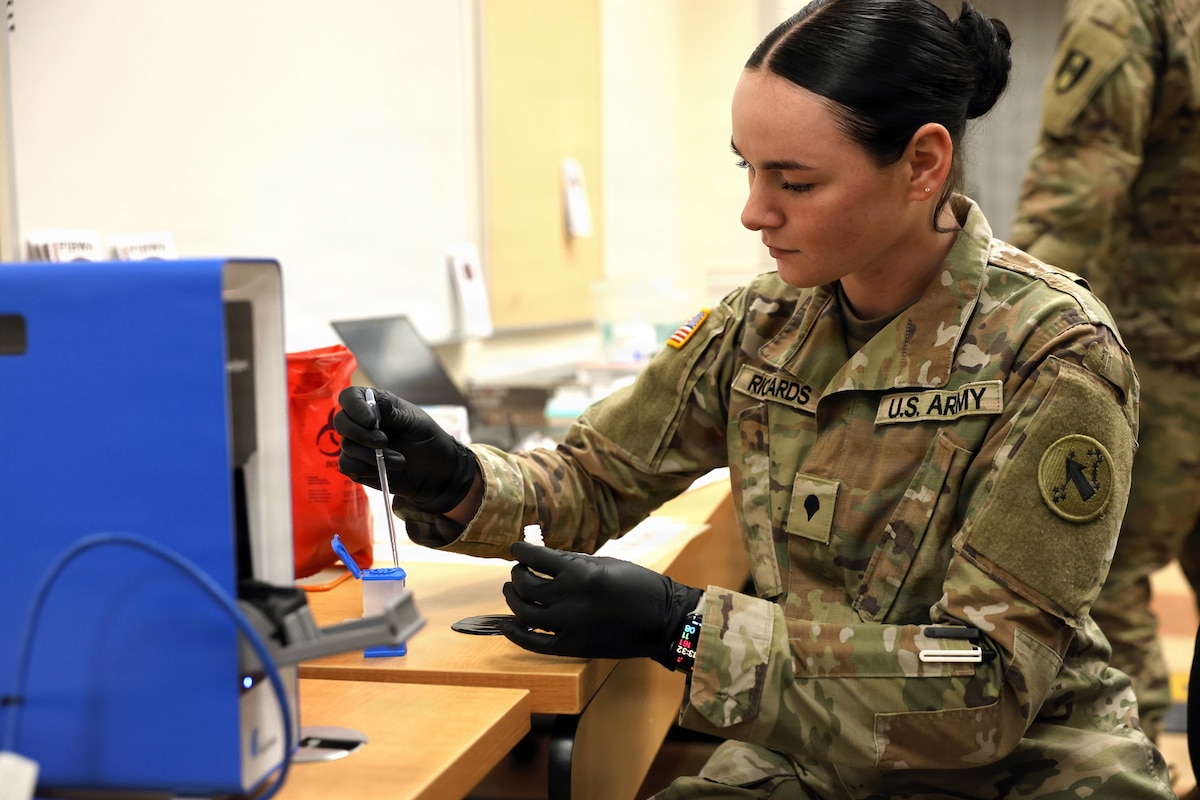 A soldier wearing black disposable gloves uses a glass dropper at a table near a blue box-like device.