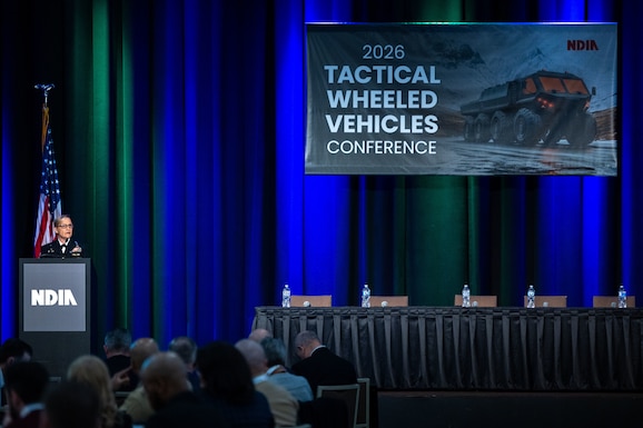 A woman speaks to an audience in a ballroom. She is wearing a Navy dress uniform.