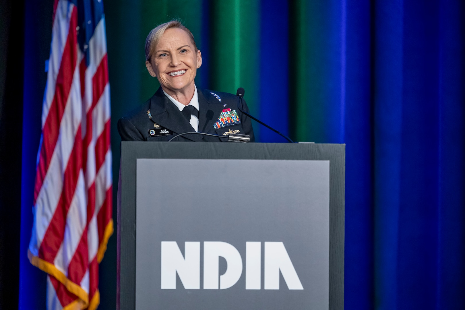 A woman in a dress Navy uniform smiles while at a podium.