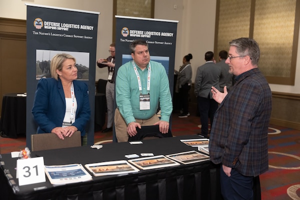 Two men and a woman have a discussion at a DLA booth in a conference setting.