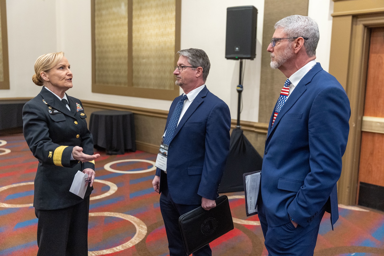 A blon haired woman in a Navy dress uniform chats with two men in suits.