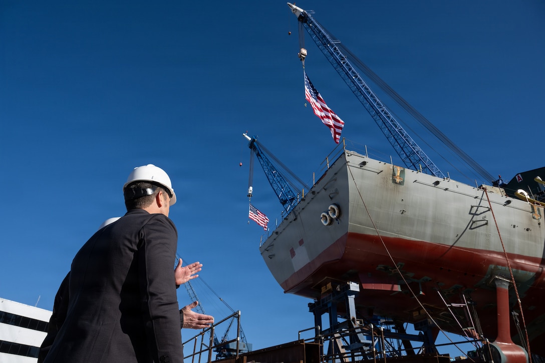 A civilian in a hard hat looks up at a boat on a metal lift outdoors.