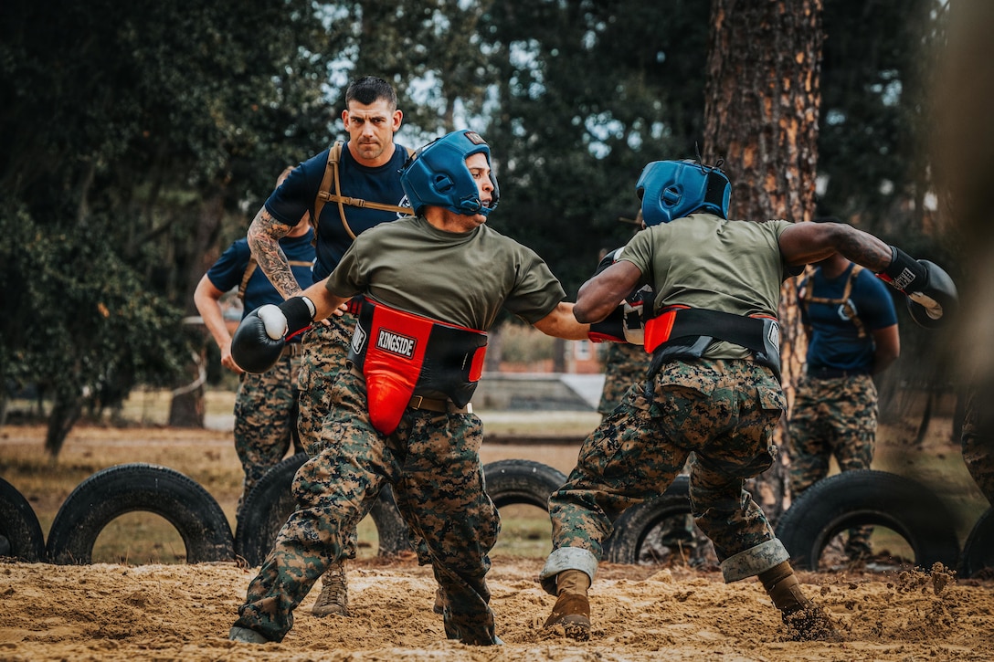 Two people wearing camouflage military uniforms and boxing headgear and gloves box in a muddy area, while another person in similar attire observes; tires and trees are in the foreground.