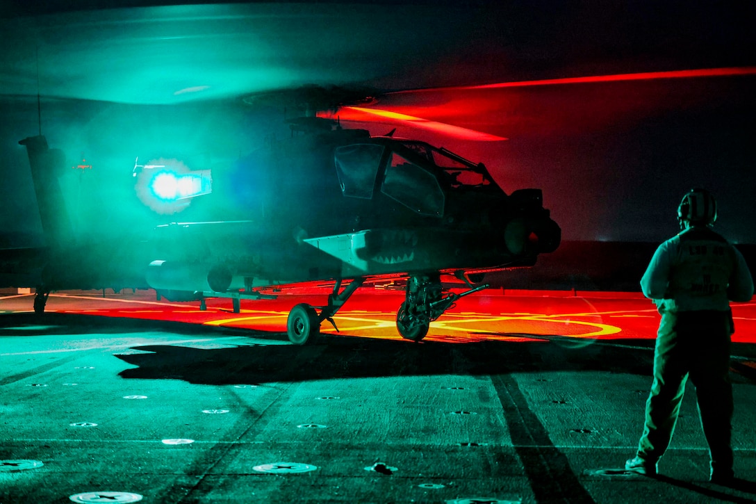 A helicopter sits on the flight deck of a ship at night, as red, green and white lights emit from the back; the silhouette of a person wearing a helmet stands in the foreground.
