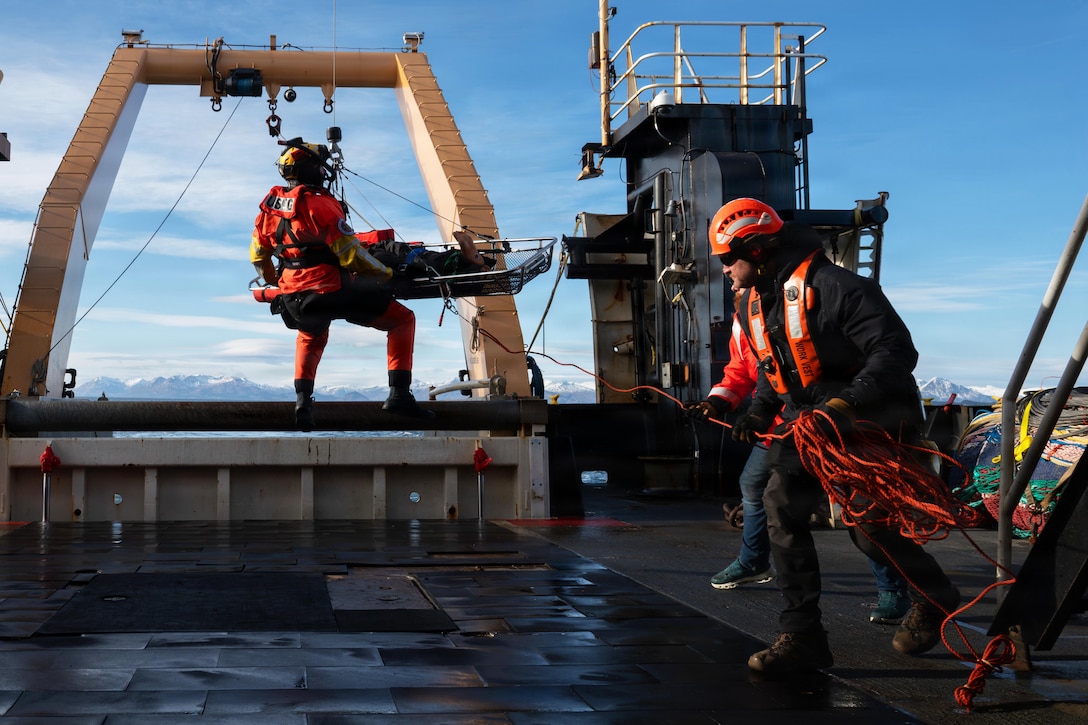 A person wearing a red vest and helmet holds a line attached to another person in similar attire, who is strapped into a harness and holding a stretcher, while being lifted into the air.