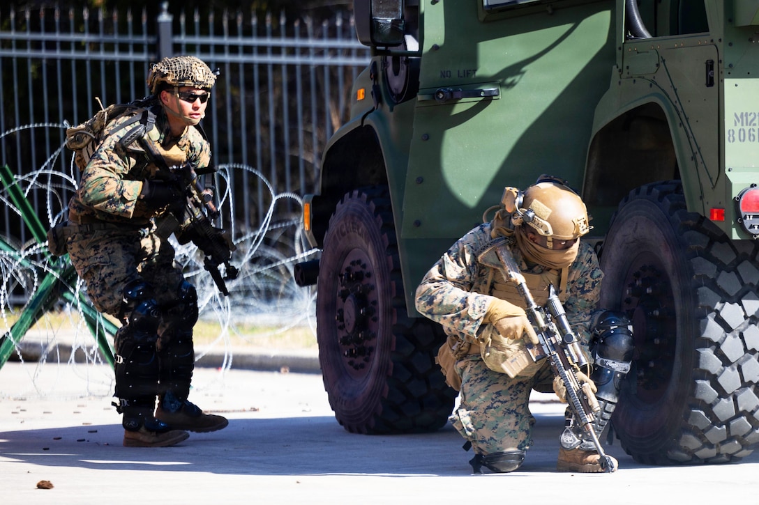 Two people wearing camouflage uniforms and helmets hold weapons and crouch near a military vehicle outside; behind them are concertina wire and a metal fence.