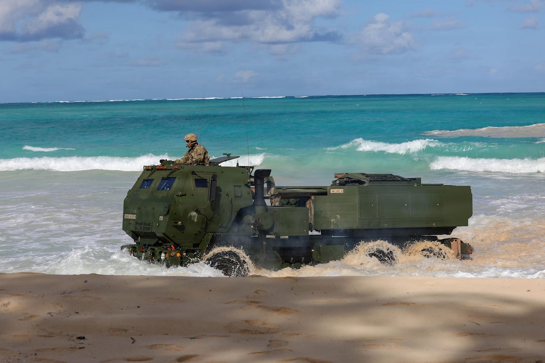 A person wearing a camouflage military uniform stands out of an open hatch of a large military vehicle moving onto a beach under a blue sky, with blue-green water in the background.