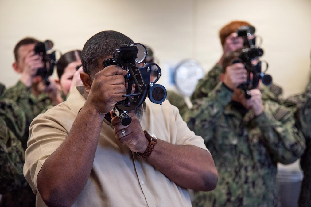 A man wearing casual attire looks through a navigational tool indoors, while other people wearing camouflage uniforms perform the same action in the background.