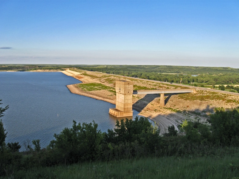 A body of water with green vegetation in the foreground, a brown concrete tower and road and green vegetation in the background.