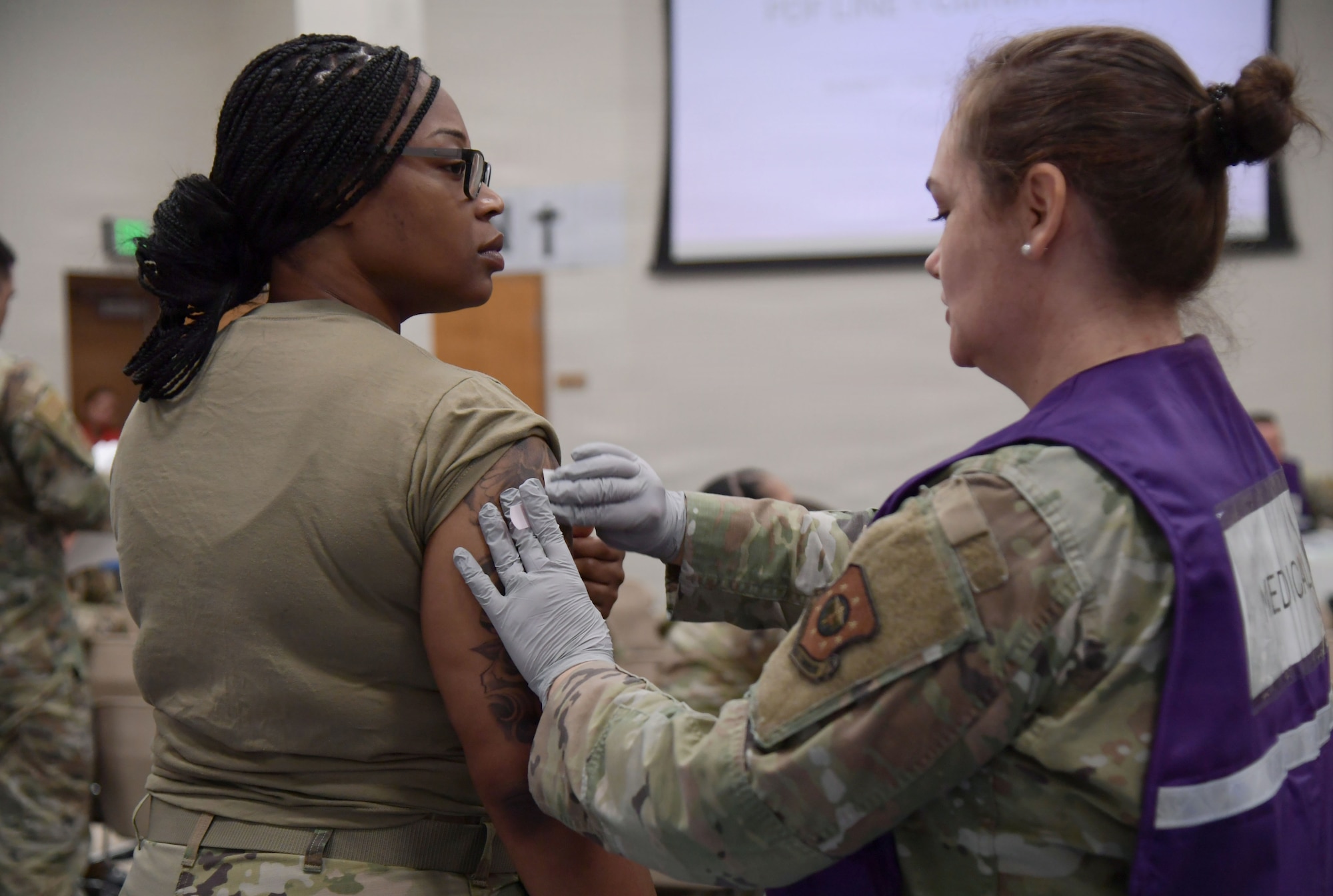A woman in a purple vest administers a shot to a woman in military uniform.