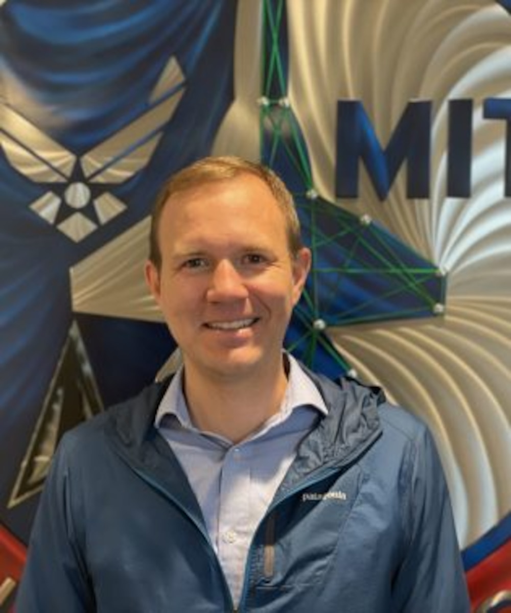 Candid photo of Air Force Lt. Col. Adam Goobic, IMA to the DAF Director of the AI Accelerator standing in front of DAF-MIT banner