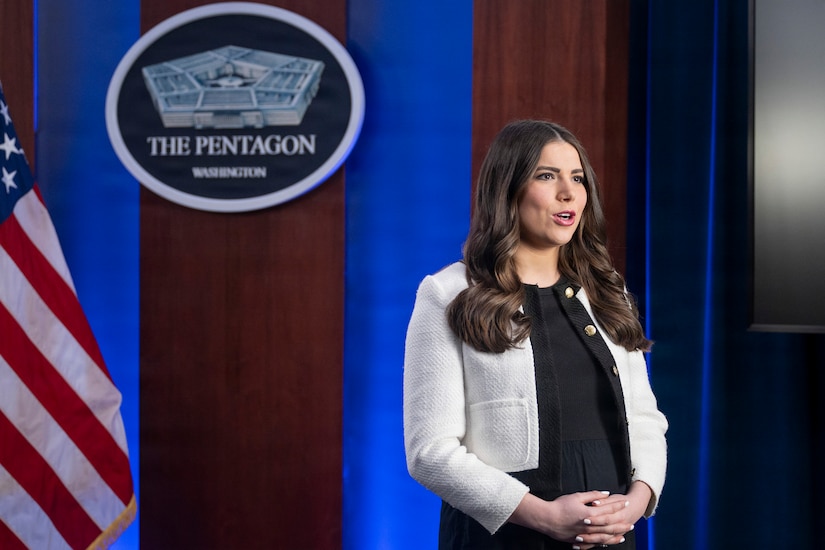 A woman in business attire stands in front of an American flag and speaks into an off-screen camera. Behind her is a sign on the wall that reads, The Pentagon, Washington.