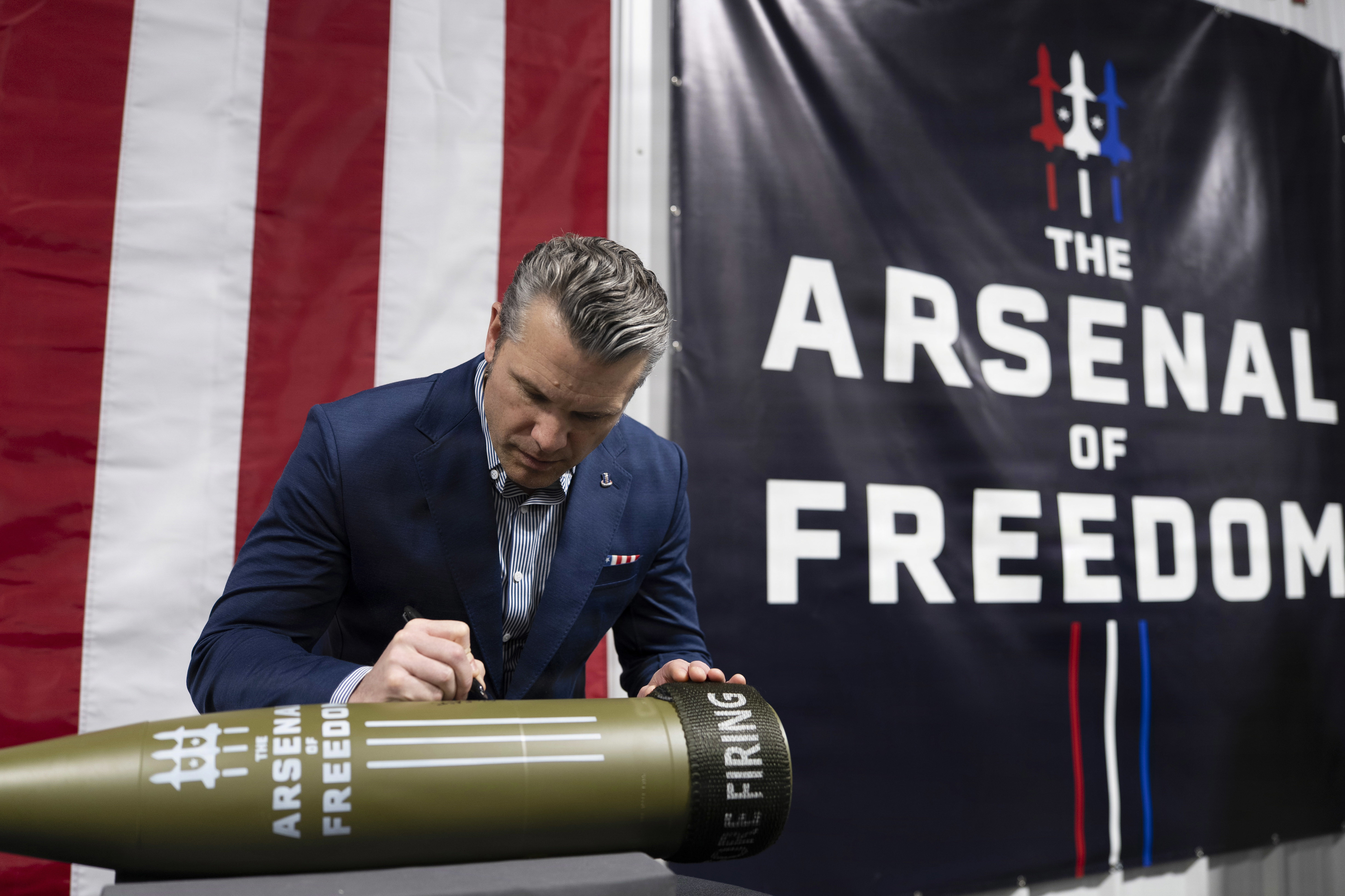 A man signs a missile in front of a flag with red and white stripes and a dark 'Arsenal of Freedom' banner in the background. 