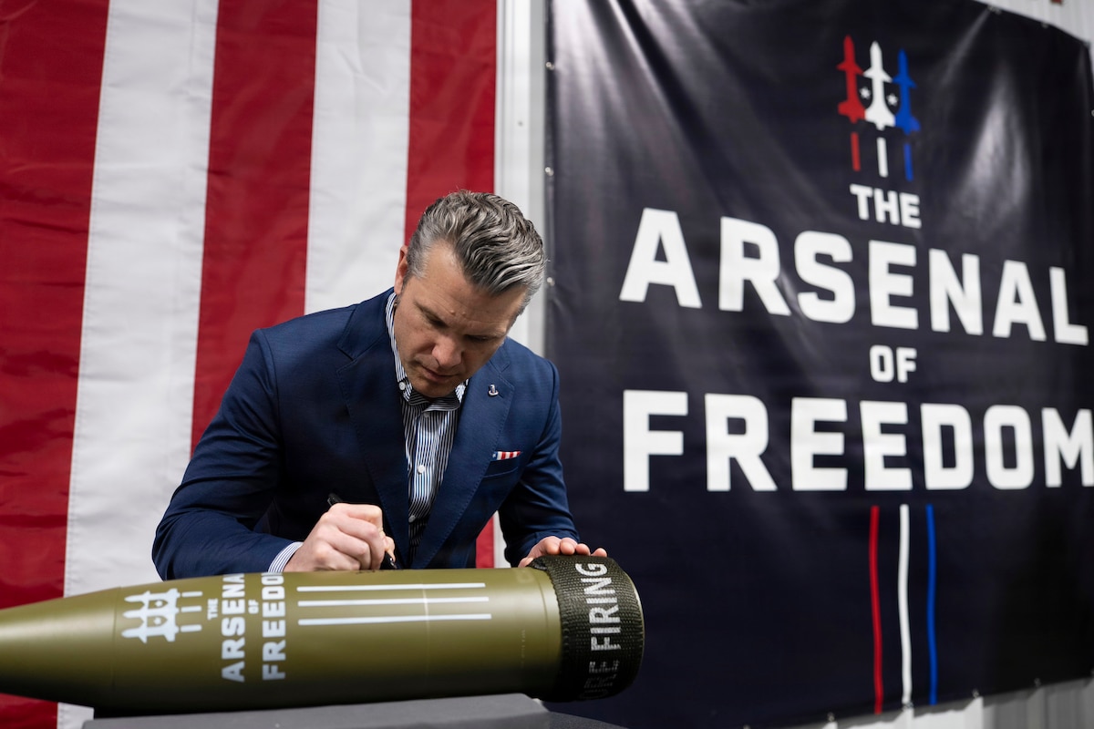 Secretary of War Pete Hegseth signs a custom missile in front of the red and white stripes and the dark Arsenal of Freedom banner in the background.