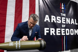 Secretary of War Pete Hegseth signs a custom missile in front of the red and white stripes and the dark Arsenal of Freedom banner in the background.