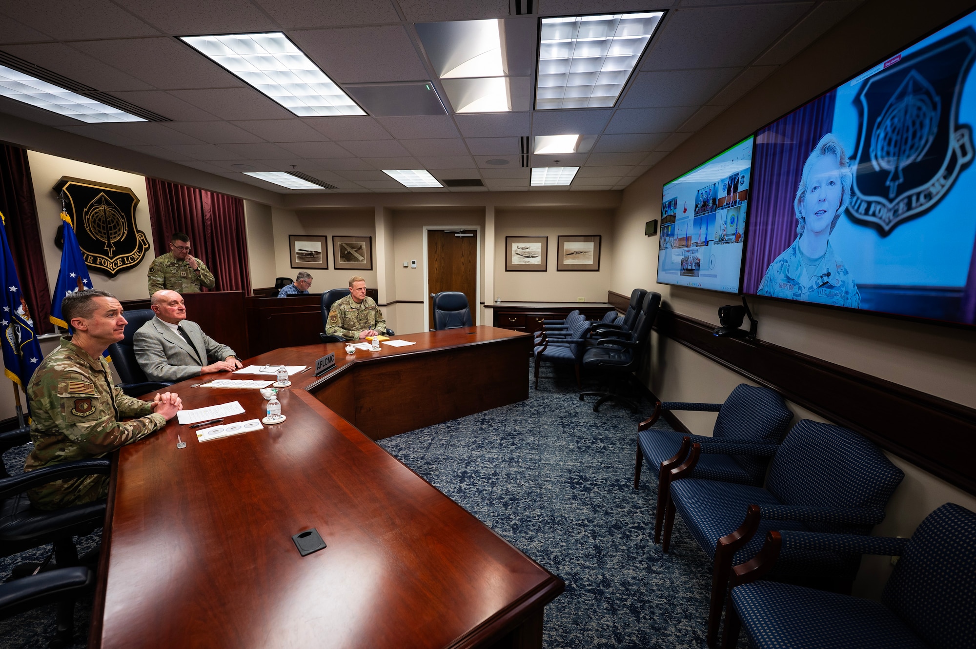 Three men sit at long table and watch a woman in Air Force camo on video screen.